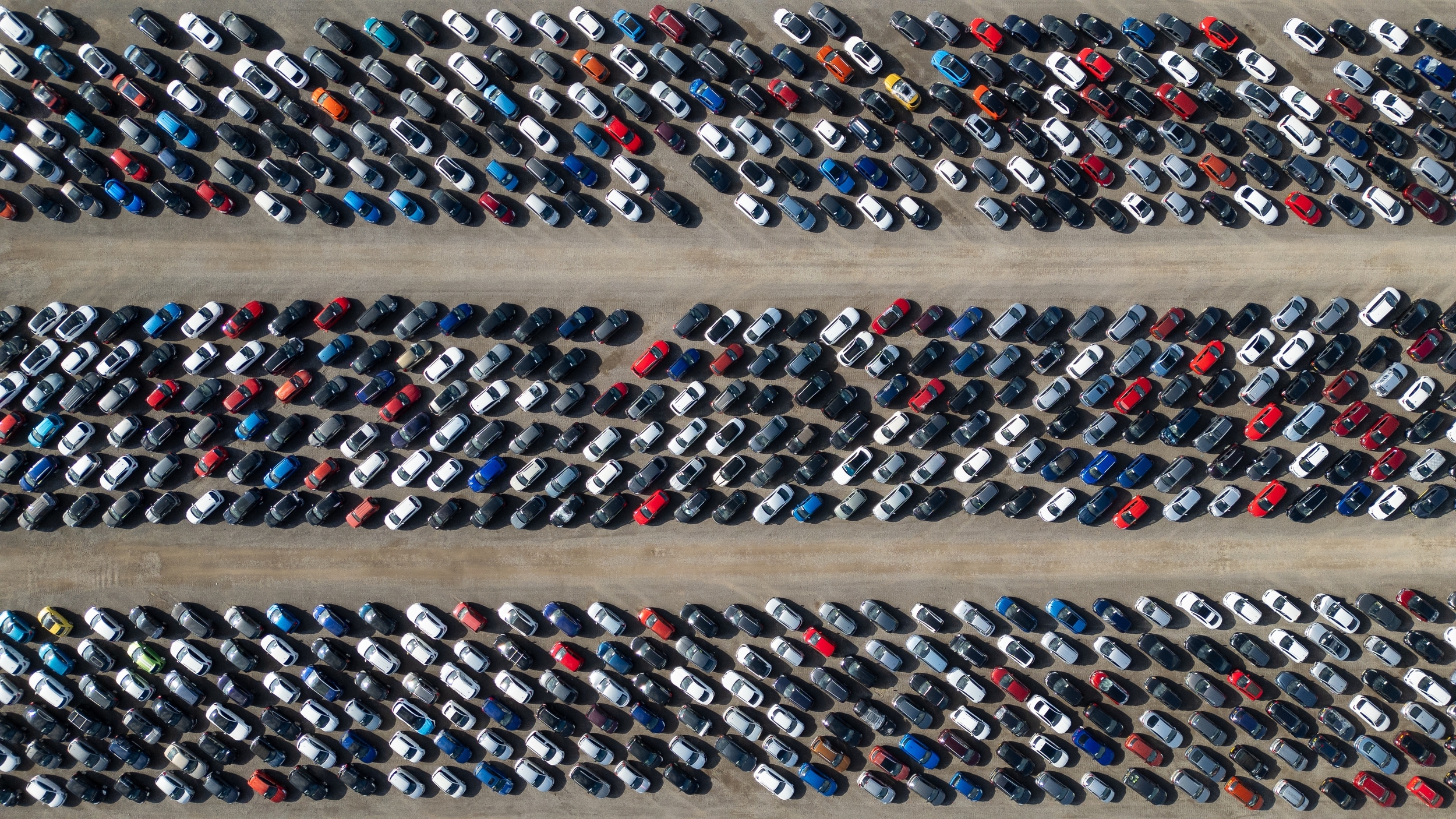 Aerial view of a large outdoor car park with hundreds of cars parked in long, diagonal rows separated by wide access lanes. Most vehicles are white, black, grey or blue, with a few red, orange and yellow cars adding contrast. The image fills the frame with repeating patterns of tightly parked vehicles, viewed from directly above.