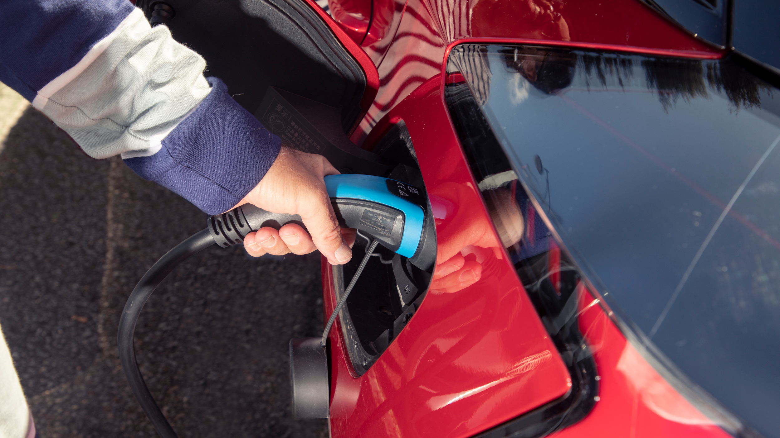 A hand plugs a charging cable into the charging port of a red electric car.