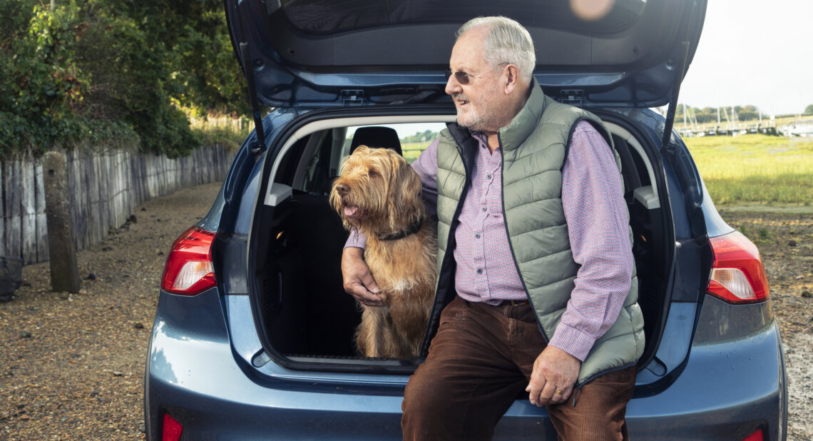 An older man sits on the boot of a blue car with his dog beside him.