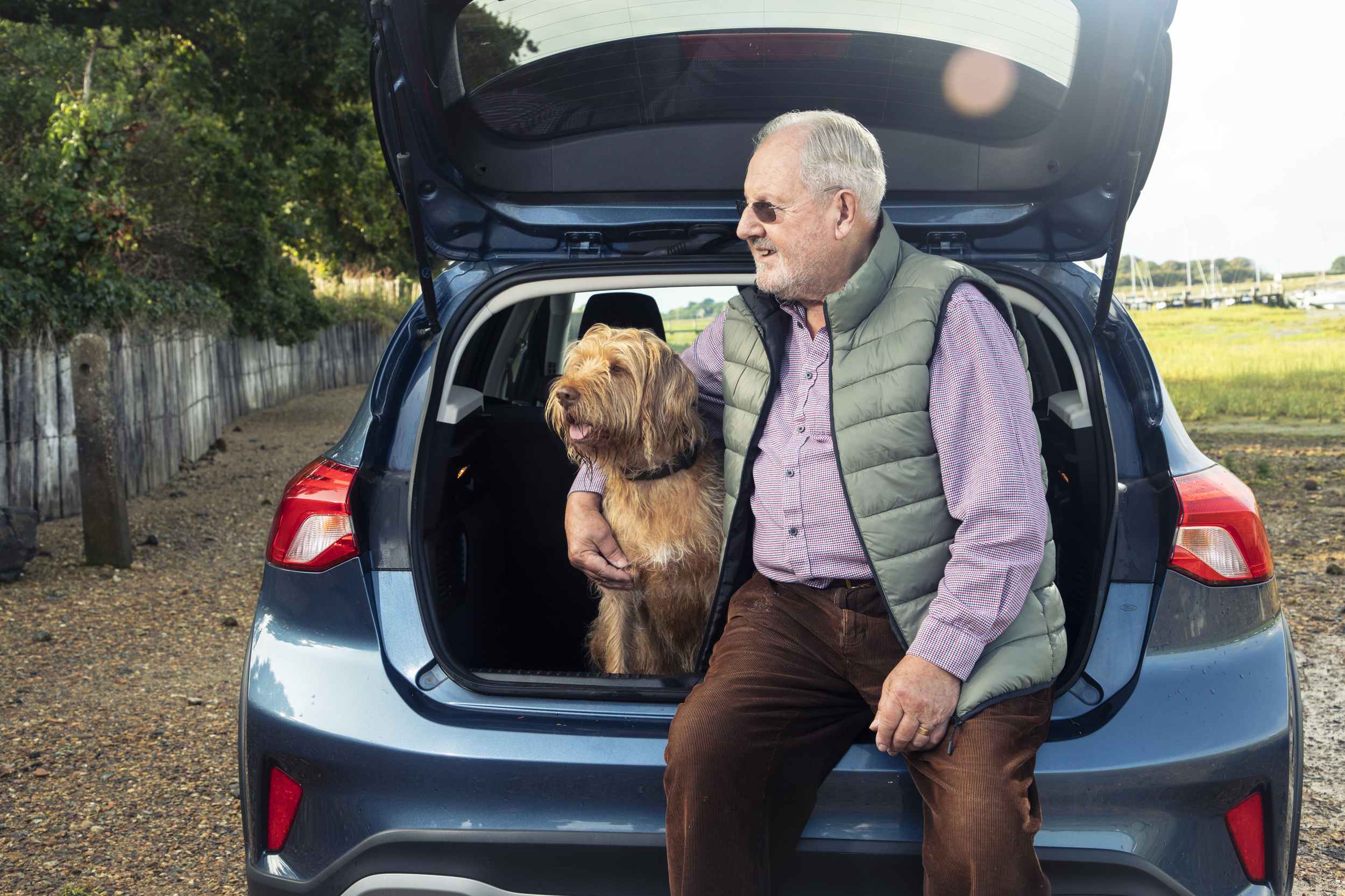 An older man sits on the boot of a blue car with his dog beside him.