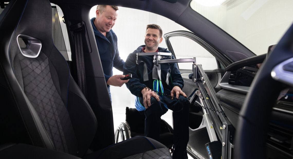 A wheelchair user tries a powered hoist that helps lift him into the passenger seat of a car, with support from another person.
