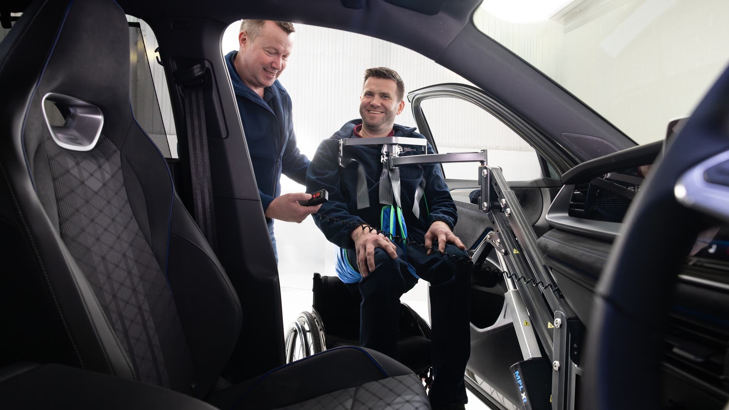 A wheelchair user tries a powered hoist that helps lift him into the passenger seat of a car, with support from another person.