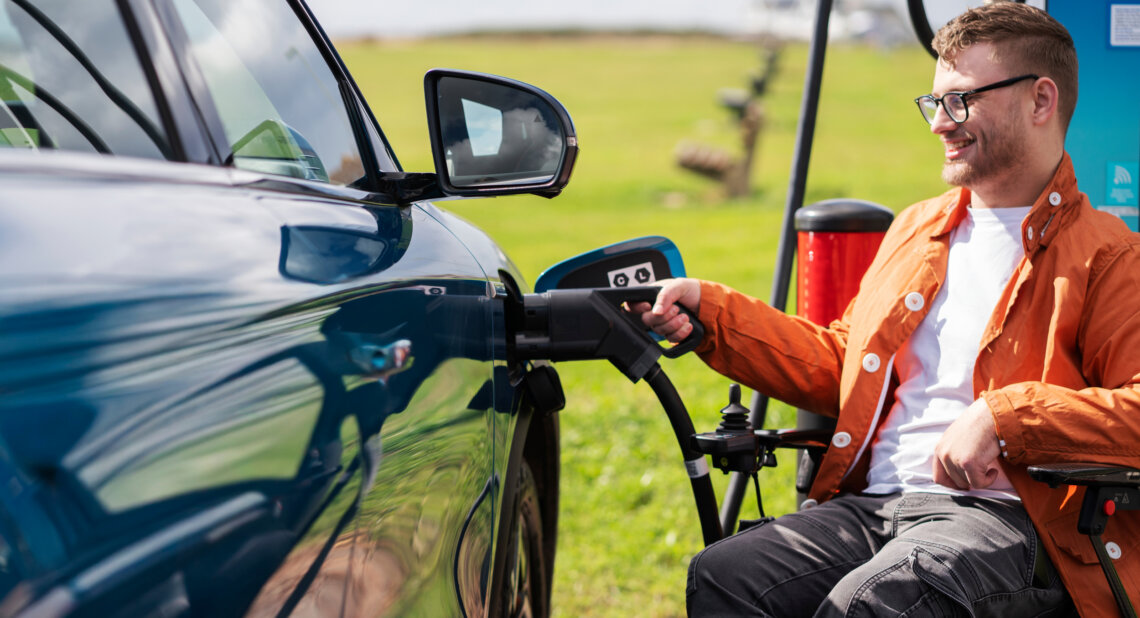 A man using a wheelchair plugs a charging cable into a blue electric Motability Scheme car at an outdoor chargepoint.