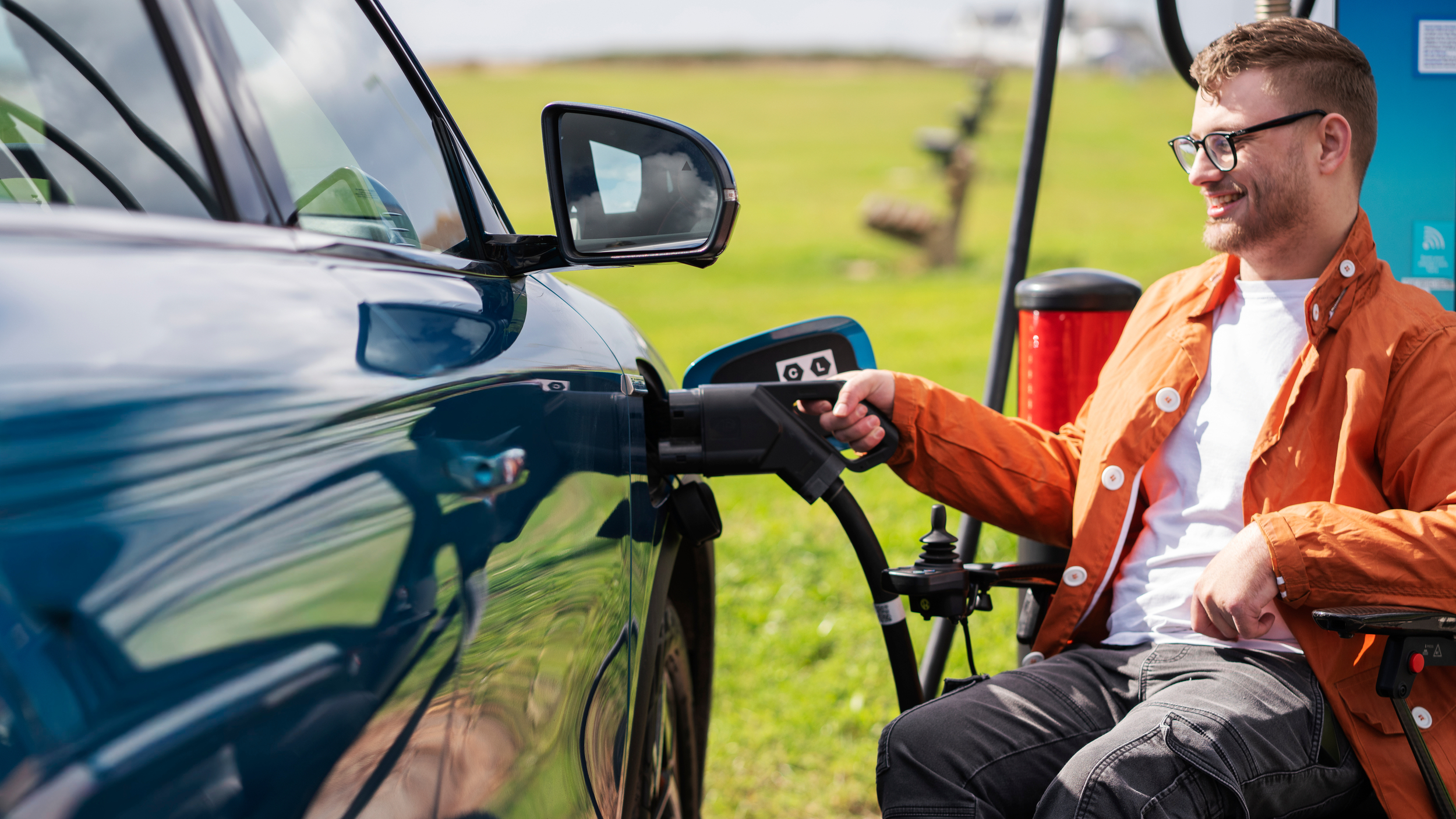 A man using a wheelchair plugs a charging cable into a blue electric Motability Scheme car at an outdoor chargepoint.